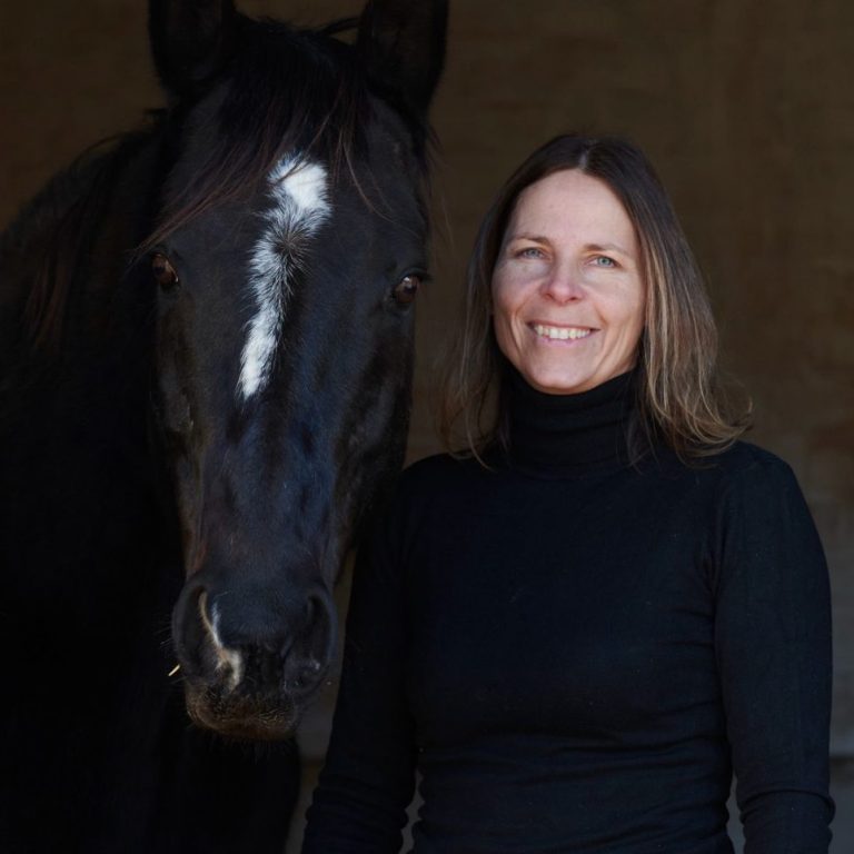 Horsebrain Coaching Denise Wiegand Denise Weigand in schwarzem Rollkragenpullover steht neben einem schwarzen Pferd mit weißen Streifen auf dem Kopf.